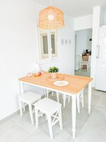 Un comedor luminoso y blanco con una mesa de madera, dos sillas blancas, una lámpara colgante de mimbre y un ventanal con marcos blancos.