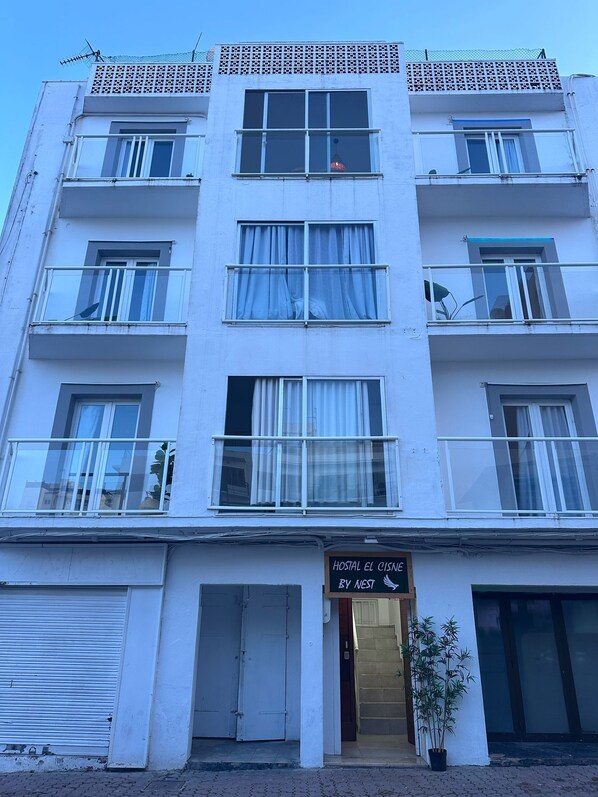 Un edificio de varios pisos con balcones, un letrero que dice "Hostal El Cisne By Nest" y una maceta con planta afuera.