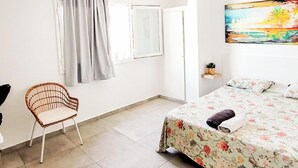 Un dormitorio con cama, una silla, ventana con cortinas, un cuadro en la pared y un pequeño estante con una planta.
