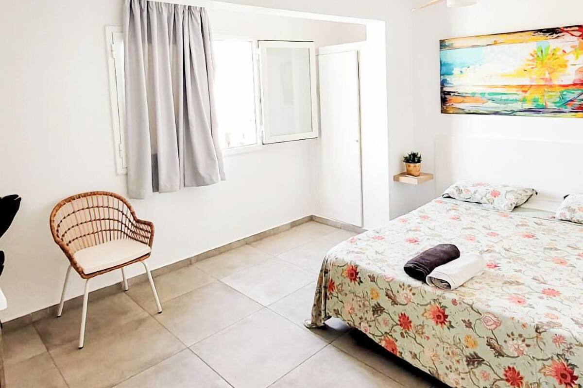 A bedroom with a bed, a chair, a window with curtains, a painting on the wall, and a small shelf with a plant.