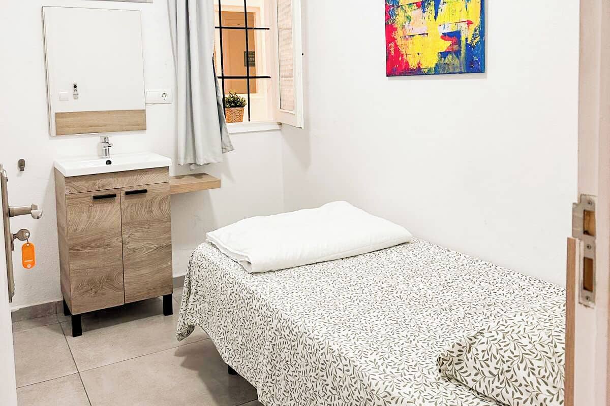 A small, clean room with a bed, a bedside table, a mirror, and a window with curtains.