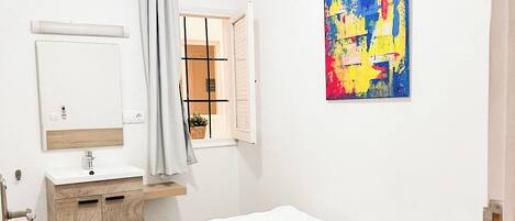 Una habitación pequeña y limpia con cama, mesita de noche, espejo y ventana con cortinas.
