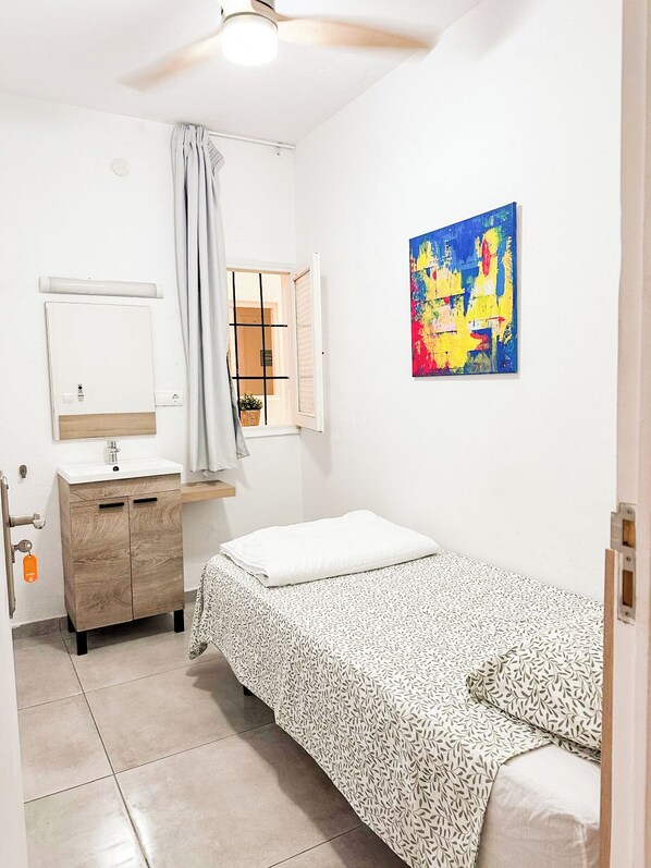 Una habitación pequeña y limpia con cama, mesita de noche, espejo y ventana con cortinas.