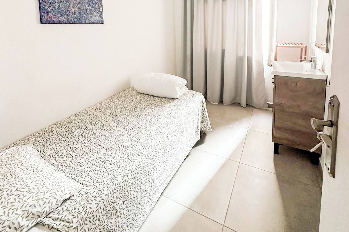 A single bed with a patterned bedspread, a ceiling fan, a painting on the wall, a bathroom with a mirror and sink, and a window with curtains.