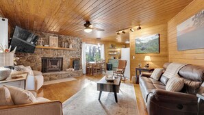 A cozy living room with a stone fireplace, wooden ceiling, and a large window overlooking a forest. - Bear Brow Cabin. Amazing view from brow to valley below. (Mentone)