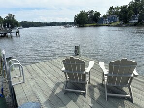 A wooden deck with two Adirondack chairs facing a calm lake, a dock with boats, and houses in the background.