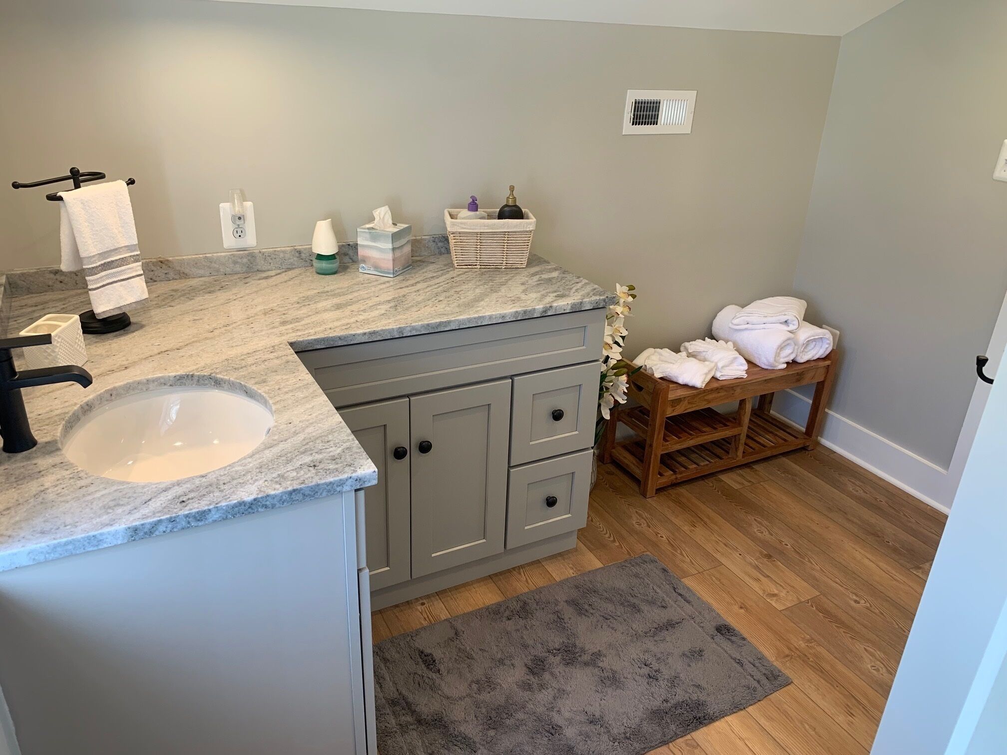 A bathroom with a marble sink, grey cabinets, a wooden bench with towels, and a grey rug.