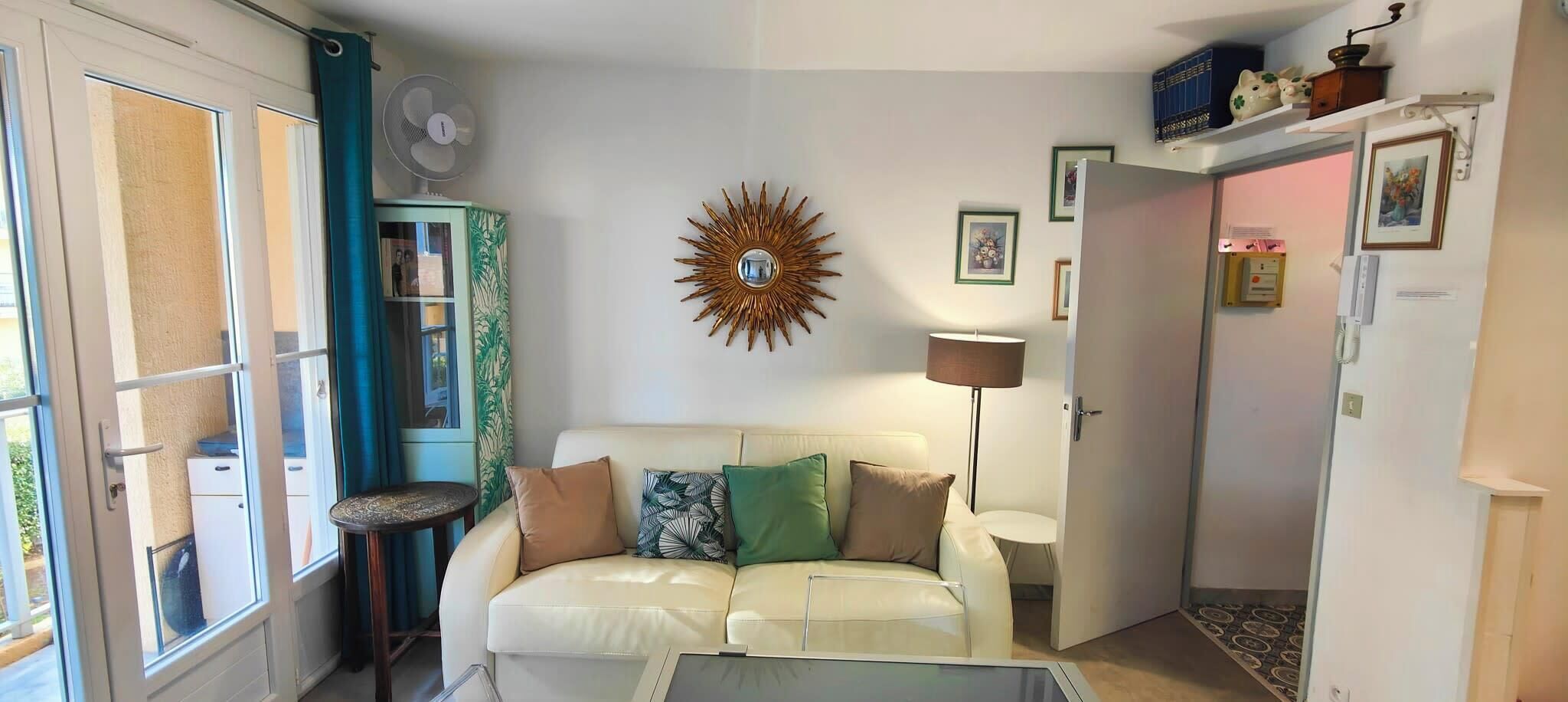 A living room with a white sofa, a small round table, a wall-mounted fan, and a decorative sunburst mirror.