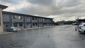 A parking lot with a row of single-story motel buildings, each with a blue door and a balcony. - Budget Motel (Orlando)