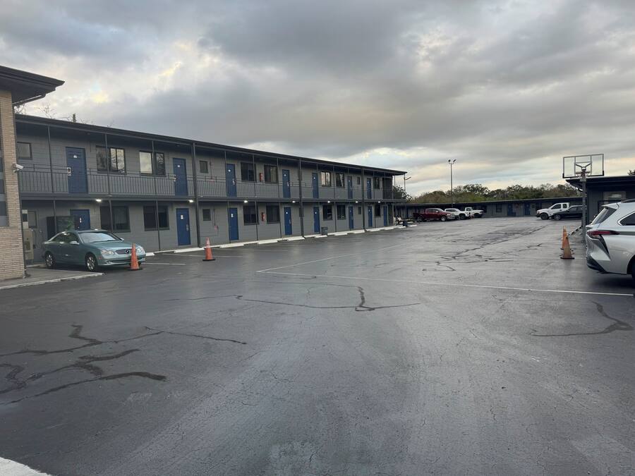 A parking lot with a row of single-story motel buildings, each with a blue door and a balcony.
