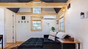 Ein gemütliches Zimmer mit einer dunklen Couch, einem Holztisch, einer Hängelampe und einem Fenster mit Jalousien.