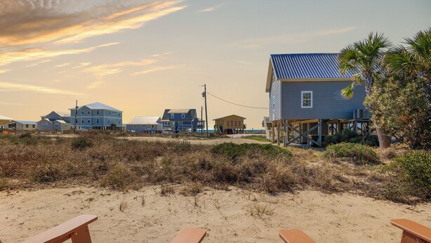 Blick auf einen Strand mit Holzliegen, Sand und Häusern mit blauen Dächern.