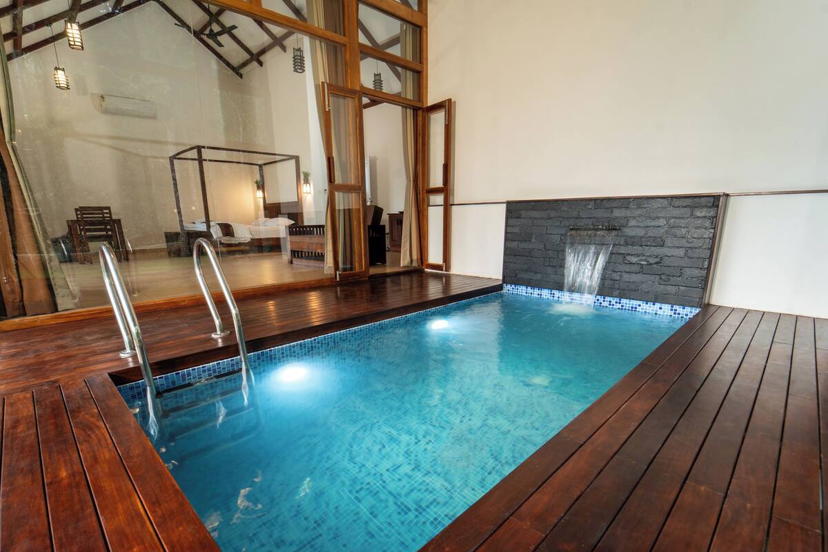 Una piscina con cubierta de madera, un muro de piedra con fuente de agua y un muro espejado que refleja la zona de la piscina.