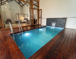 Una piscina con cubierta de madera, un muro de piedra con fuente de agua y un muro espejado que refleja la zona de la piscina.