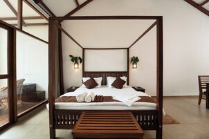 Una cama con dosel de madera, ropa blanca de cama y almohadas marrones. Hay dos mesitas de noche con lámparas y una silla a la izquierda.