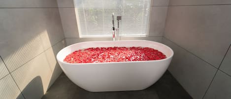 Un baño moderno con una bañera independiente llena de pétalos de rosa, una ventana con persianas y paredes de azulejos.