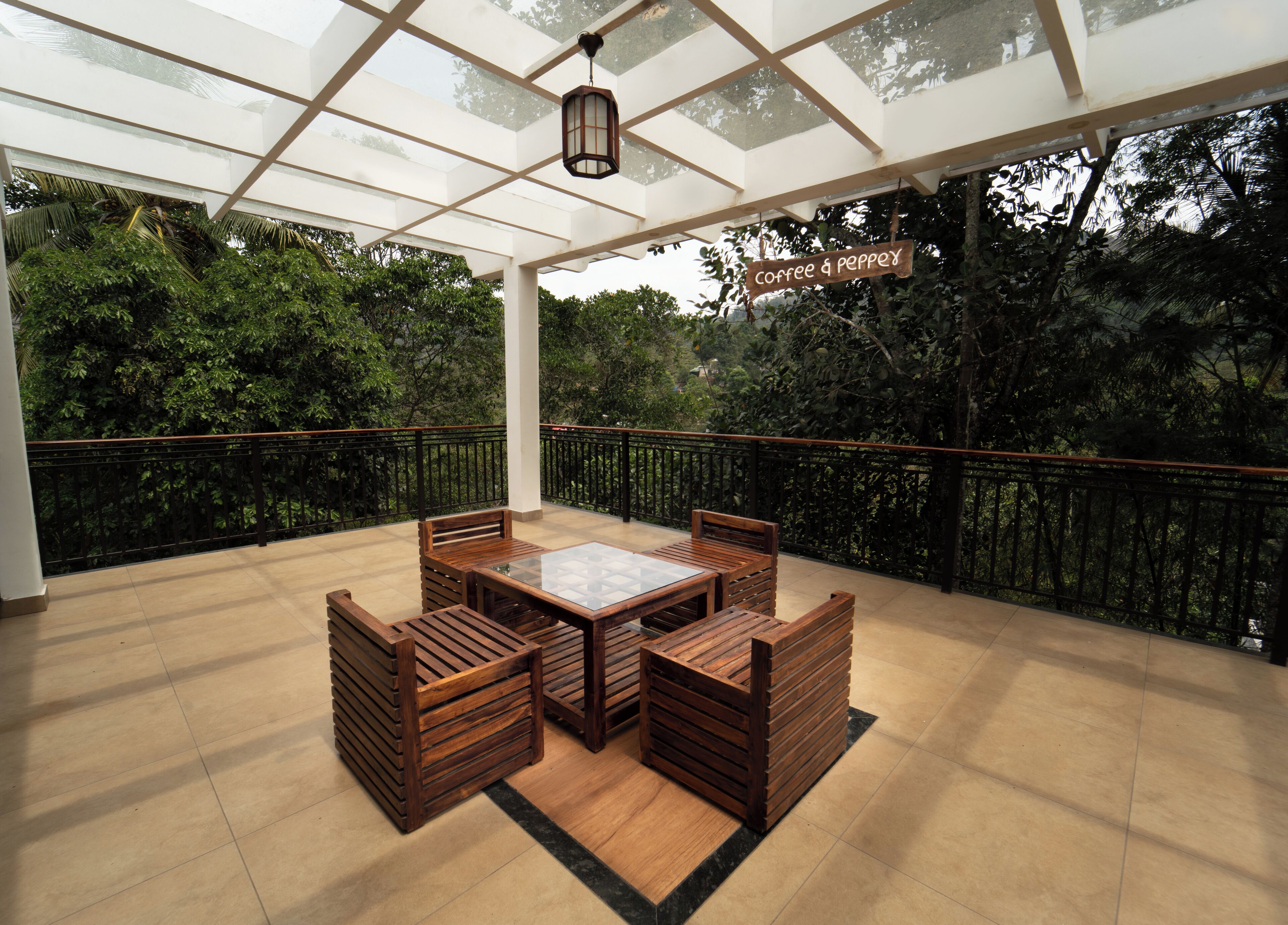 A covered patio with a wooden table and chairs, surrounded by a glass railing, overlooking a lush garden.