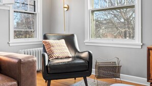 Un salón moderno con un sillón de cuero negro, un cojín estampado, una lámpara de pie y vistas a árboles a través de amplios ventanales.
