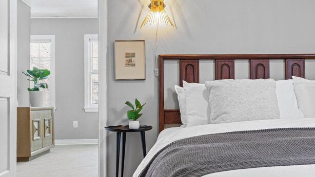 Un dormitorio con cabecera de madera, ropa de cama blanca, una mesita de noche con una planta y una lámpara de pared.