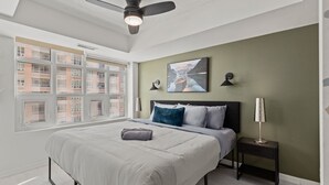Ein Schlafzimmer mit einem Bett, einem Deckenventilator, einem Nachttisch, einer Lampe und einem Fenster mit Blick auf Gebäude.