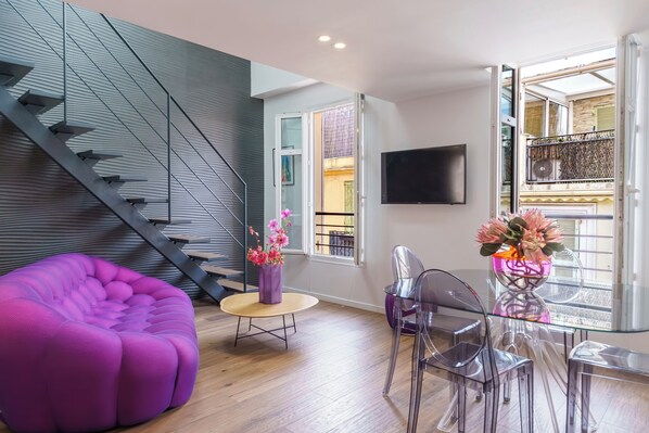 A modern living space with a staircase, a purple sofa, a glass dining table with chairs, and a TV mounted on the wall.