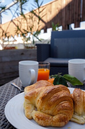 Un desayuno con cruasanes en un plato, un vaso de zumo de naranja y una taza, con una valla de madera y mobiliario exterior al fondo.