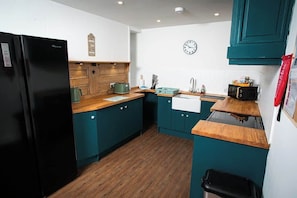 Una cocina compacta con gabinetes verde azulado, un refrigerador negro, encimeras de madera, un reloj de pared y un pequeño cubo de basura.