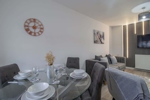A modern living room with a dining area, a large wall clock, a flat-screen TV, and a sofa.