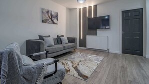 A modern living room with a grey sofa, a patterned rug, a mounted TV, and a door. - The Larky - Modern 2 Bed Cottage in Larkhall (Larkhall)