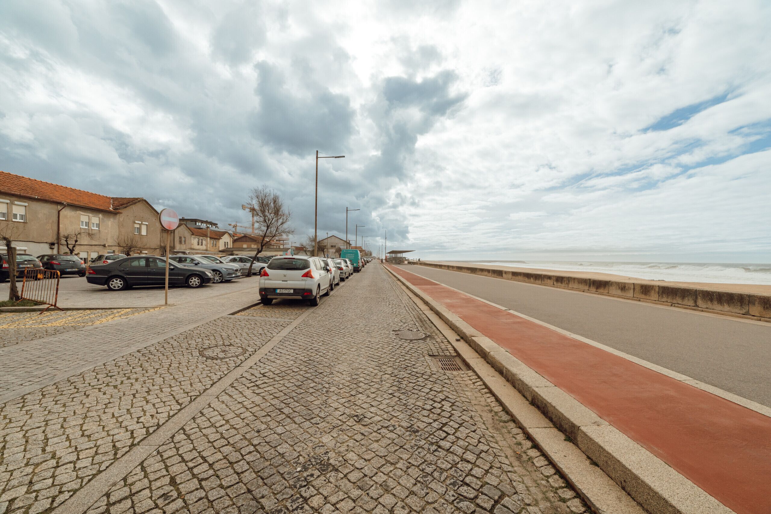 A cobblestone street lined with parked cars and houses, leading to a beach.