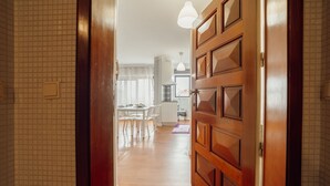 Un couloir mène à une salle à manger avec une table et des chaises. Au fond, on aperçoit une cuisine, et une porte en bois avec des panneaux de verre ferme l’ensemble.