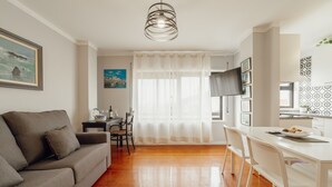 Un salon moderne avec un canapé gris, une table à manger avec des chaises blanches, un téléviseur et un coin cuisine avec des carreaux décoratifs.