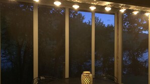Uma área de estar coberta ao ar livre, com mesa e cadeiras de metal redondas, um candeeiro decorativo e grandes janelas com vista para árvores.