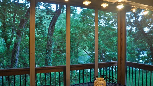Um deck de madeira com mesa e cadeiras, grandes janelas com vista para uma floresta e luzes de fada.