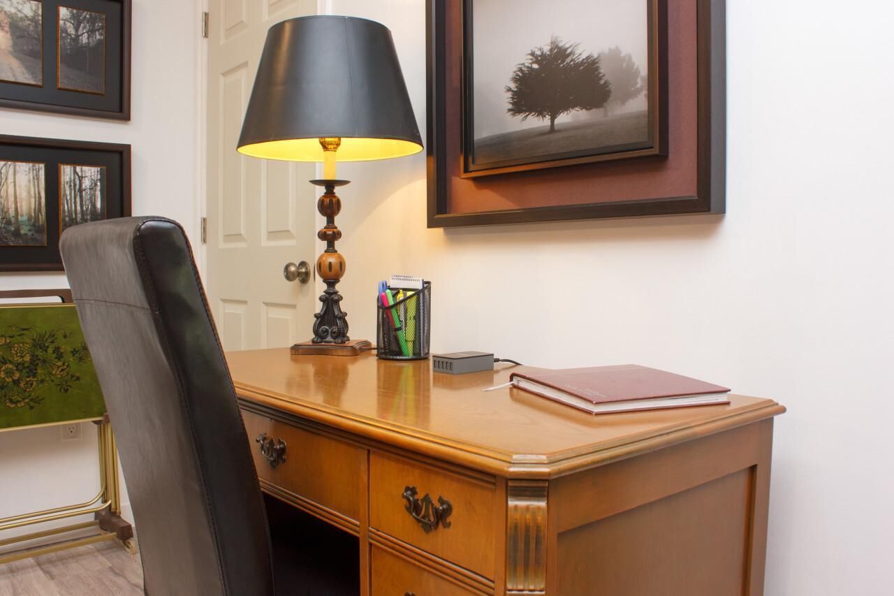 A wooden desk with a lamp, a chair, and a framed picture on the wall.