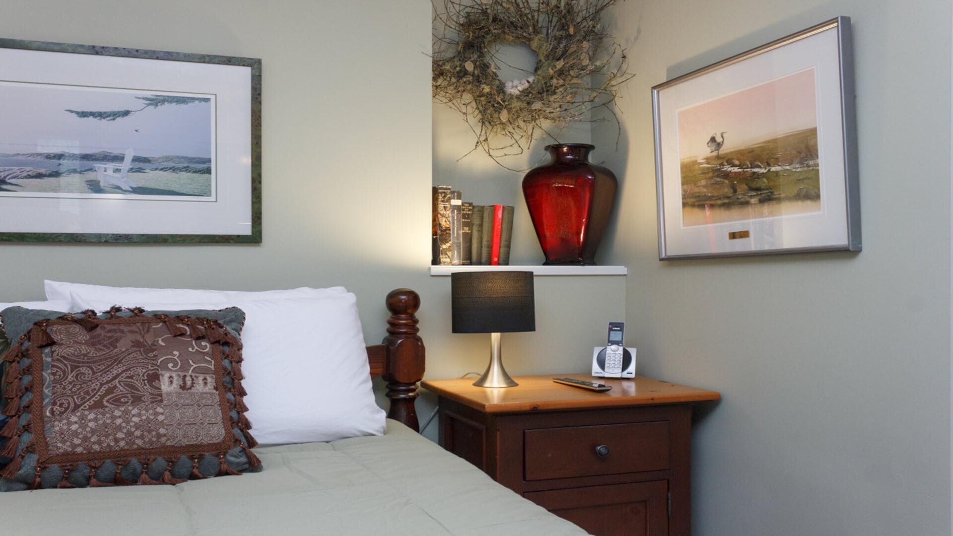 Un dormitorio ordenado con una cama, una mesita de noche con lámpara, un teléfono y una estantería con libros y un florero.