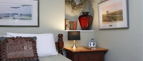 Un dormitorio ordenado con una cama, una mesita de noche con lámpara, un teléfono y una estantería con libros y un florero.