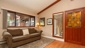 Una sala de estar con un sofá marrón, un tapete estampado, piso de madera, una puerta de madera con un panel de vitral y una ventana con persianas.