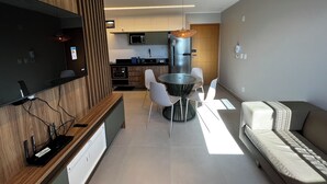 Ein modernes Wohnzimmer mit einer Couch, einem Esstisch mit Stühlen, einer Küchenzeile mit Geräten und einem an der Wand montierten Fernseher.