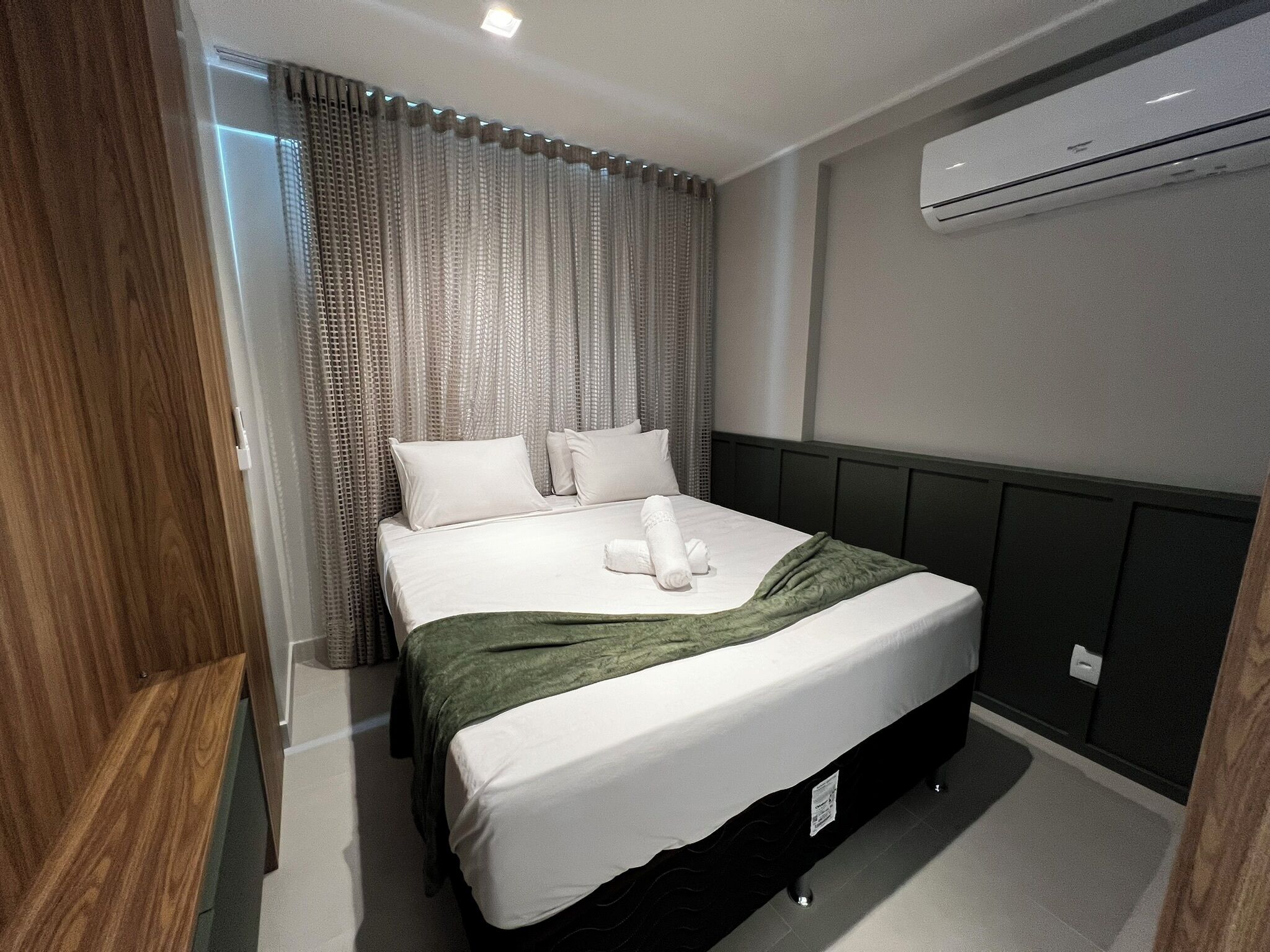 A modern hotel room with a neatly made bed, a large window with curtains, an air conditioning unit, and a wooden panel on one wall.