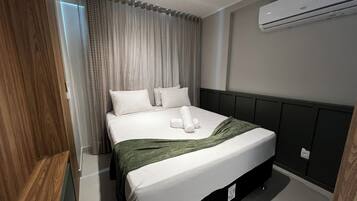 A modern hotel room with a neatly made bed, a large window with curtains, an air conditioning unit, and a wooden panel on one wall.