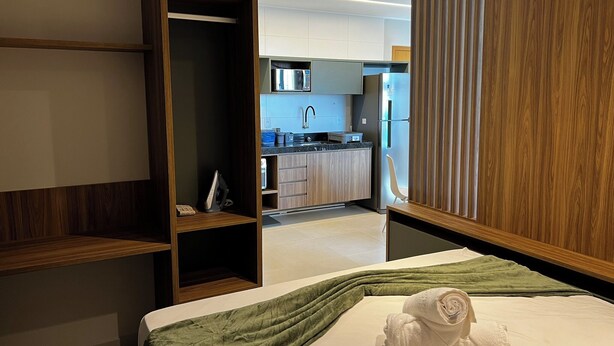 Ein modernes Hotelzimmer mit einem Bett, einer grünen Tagesdecke und einem ordentlich gefalteten Handtuch. Im Hintergrund ist ein Holfischschrank und ein Blick in den Küchenbereich zu sehen.
