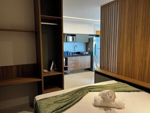 A modern hotel room with a bed, a green throw blanket, and a neatly folded towel. There is a wooden wardrobe and a view of the kitchen area in the background. - Studio Design 3 Apt 604  (Maceio)