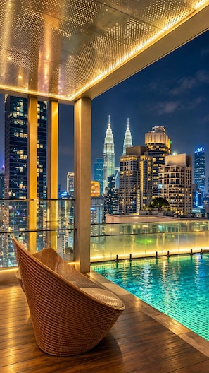 Un balcón con vista al horizonte urbano, incluyendo las Torres Petronas, una piscina y una silla de mimbre.