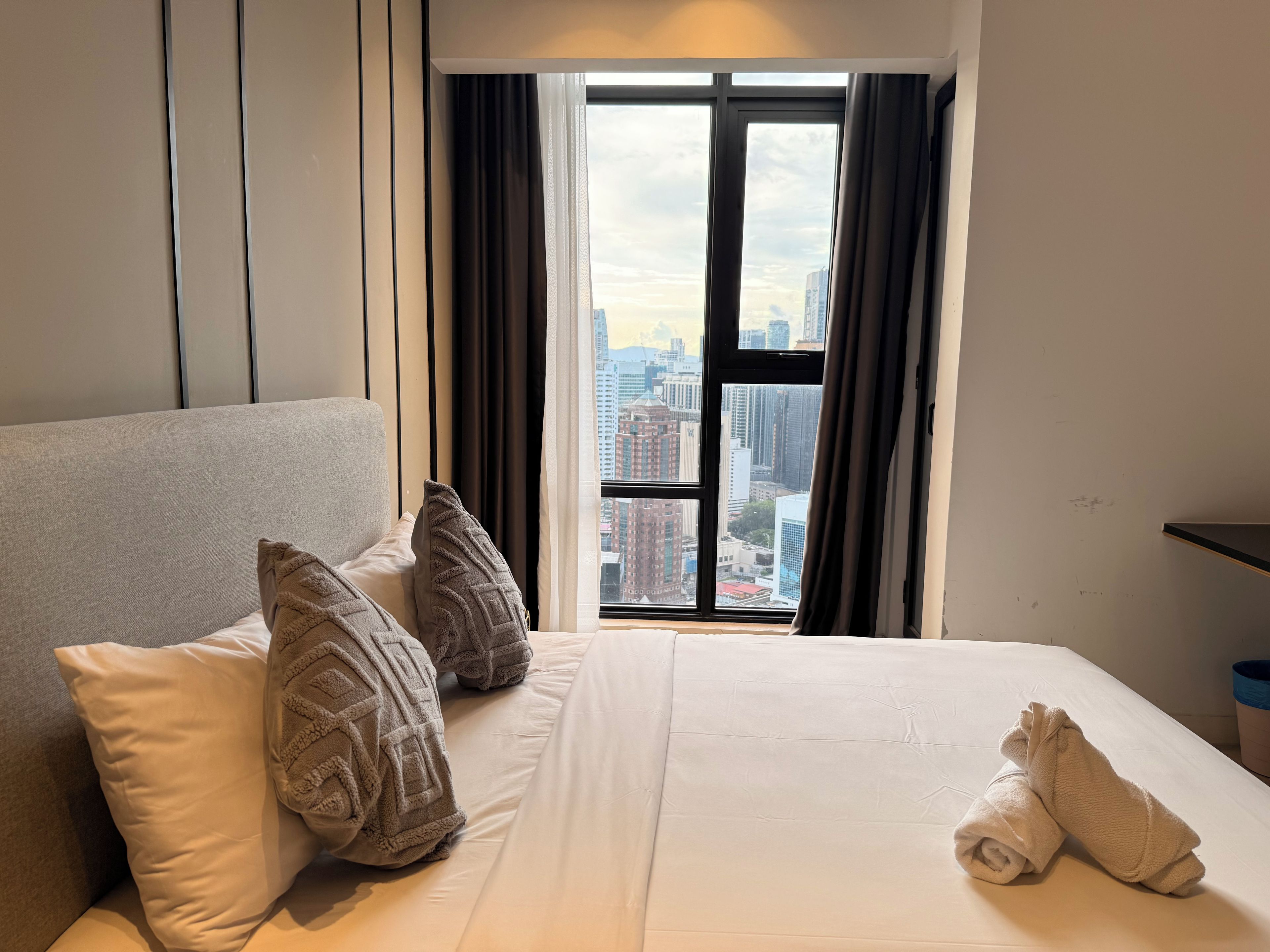 A hotel room with a large bed, two pillows, a neatly made bed, a window with curtains, and a view of the cityscape.