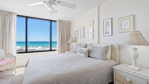 Une chambre avec une grande fenêtre donnant sur une plage, un lit recouvert d’une courtepointe matelassée, une table de chevet avec une lampe et un fauteuil.