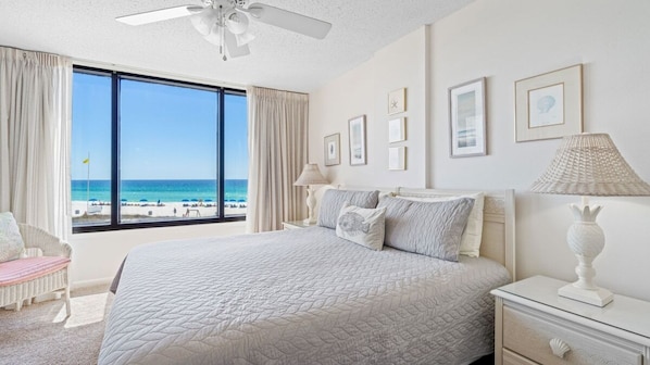 A bedroom with a large window overlooking a beach, a bed with a quilted bedspread, a nightstand with a lamp, and a chair.