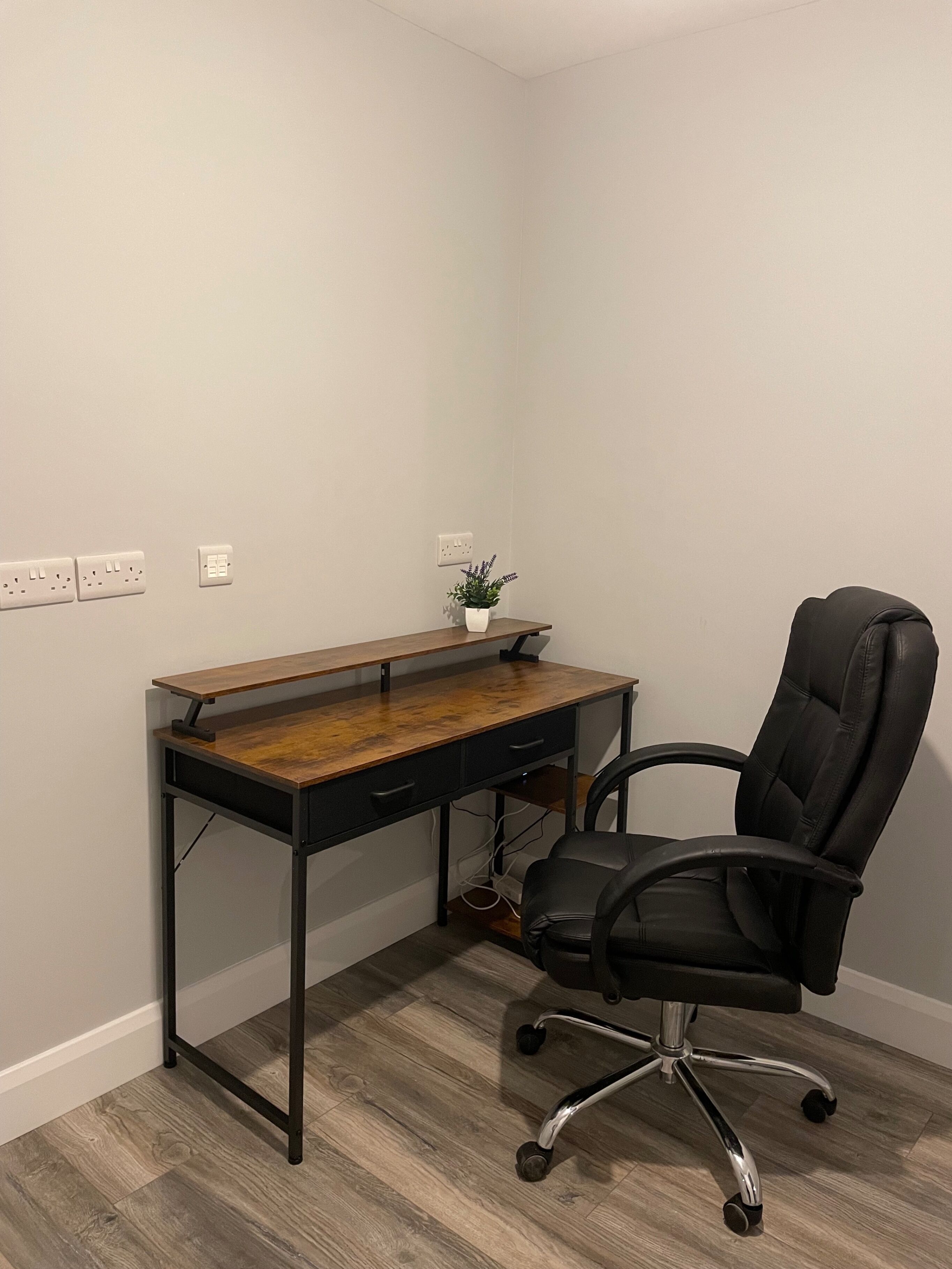 Oficina en esquina con un escritorio de madera, una silla de oficina negra, una planta en maceta y un enchufe de pared.