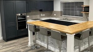 A modern kitchen with dark cabinets, a wooden island with bar stools, a built-in oven, and a window with blinds. - Spacious 5Br Home - Country View - Close to Galway (Tuam)