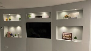 A living room with a flat-screen TV mounted on the wall, surrounded by shelves with decorative items and books. - Spacious 5Br Home - Country View - Close to Galway (Tuam)